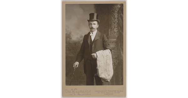 A black-and-white studio photograph of a White man with a mustache, wearing a top hat and three-piece suit, holding a cane in one hand, white cloth draped over the other arm.