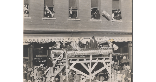 A black-and-white photograph of a group of people standing on an elevated stage in front of a building, addressing a crowd of people below.