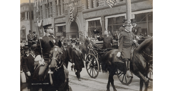 A black-and-white photograph of a parade of people on horseback, with a White man in the center in a carriage tipping his hat to onlookers.