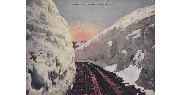 A color postcard photograph of railroad tracks curving through tall walls of snow on either side; a mountain peeking over the snow in the distance.