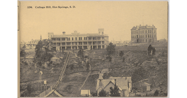 A sepia-toned photograph of, in the foreground, a three-story house with wide verandas on each level on a hill and several multi-story buildings in the background.