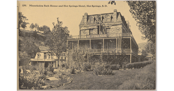 A sepia-toned postcard of a three-story stone hotel with a wrap-around porch nestled in a lush landscape.