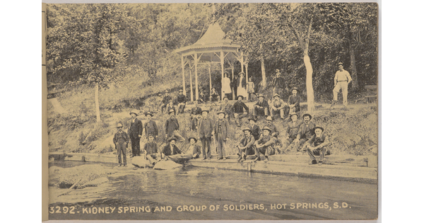A sepia-toned photograph of a group of men and women seated on a slope and in a gazebo next to a canal.