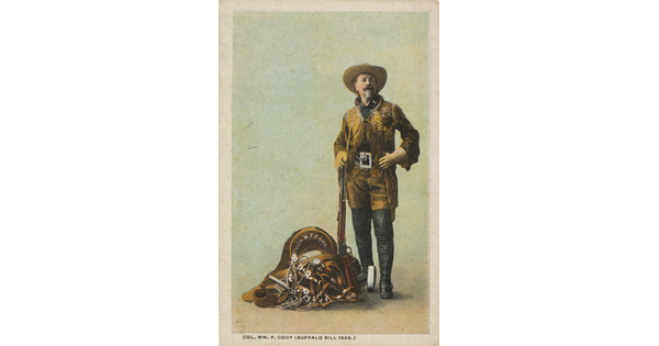 A color photograph postcard of a White man wearing a cowboy hat, brown leather jacket with fringe, tall black boots over his pants posing with a rifle and a pile of equestrian equipment.