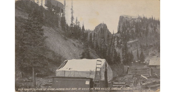 A sepia-toned postcard of a small building with a white tarp as the roof at the foot of steep mountains.