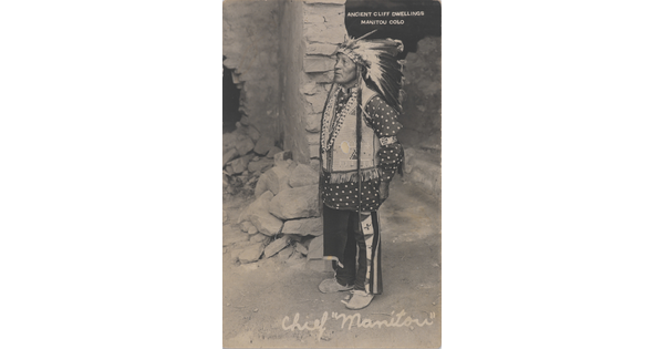 A black-and-white photograph of a Indigenous man in ceremonial dress standing in front of stone dwelling.