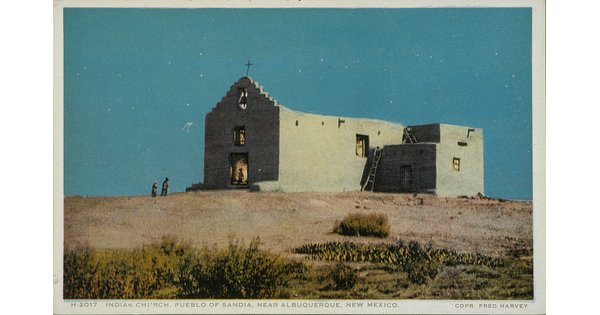A color postcard of an adobe-style church in a desert landscape with two people walking toward its open doors.