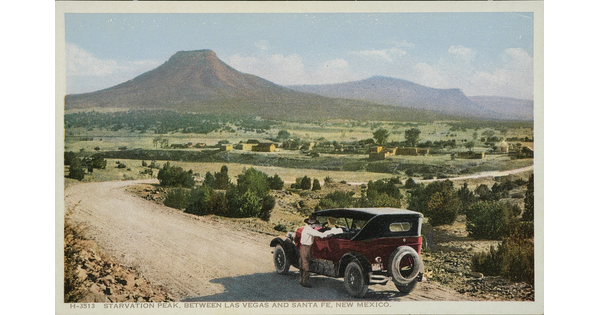 A color postcard of a man standing next to a red 1920s-style automobile on a winding dirt road with mountains in the distance.