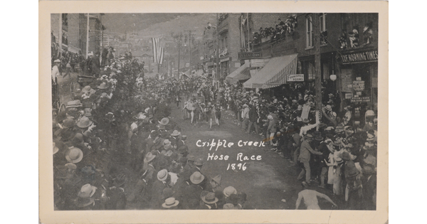 A black-and-white postcard of crowds of people lining a town street watching people harnessed to carriages running.