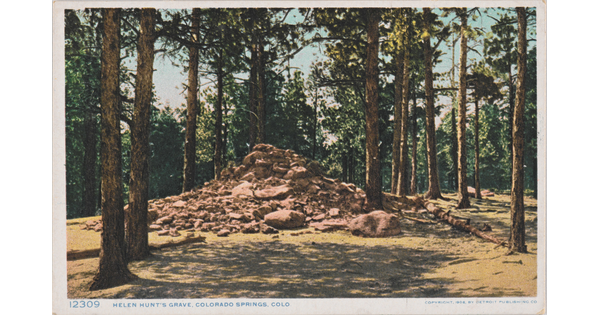 A colored photograph on a postcard of a large pile of rocks and boulders surrounded by tall pine trees.