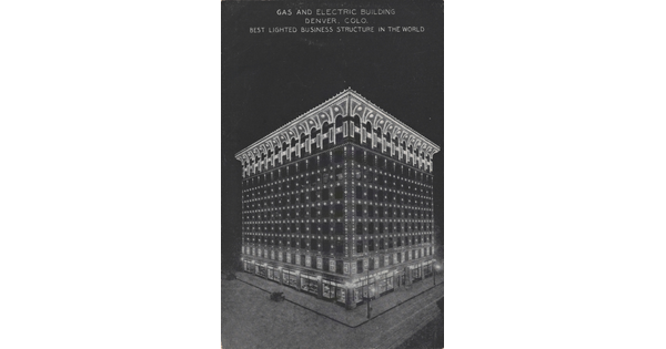 A color postcard of a nine-story building lit up at night under a full moon that is partially hidden behind clouds.