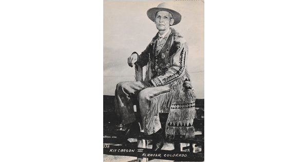 A black-and-white postcard of an older White man in western dress seated on a fence.