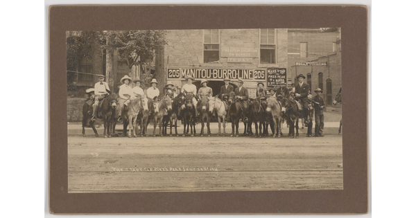 A sepia-toned photograph of a group of people, most with cowboy hats, on horses or donkeys on a dirt road in front of a brick building.
