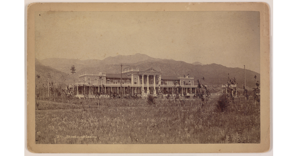 A sepia-toned photograph of a large, multi-story ornate building and mountains in the background.
