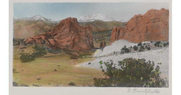A color photograph of a plain at the foot of tall, red rock formations and snowy mountains in the distance.