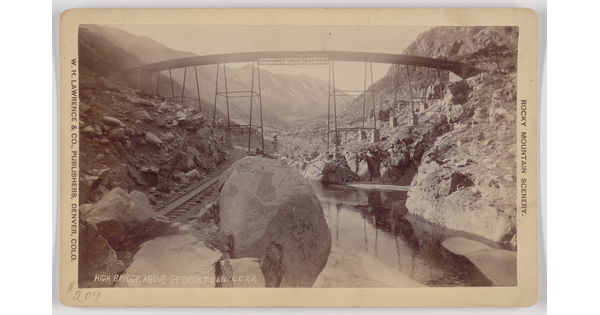 A black-and-white photograph postcard of a very tall simple truss bridge spanning railroad tracks and a deep rocky valley and river.