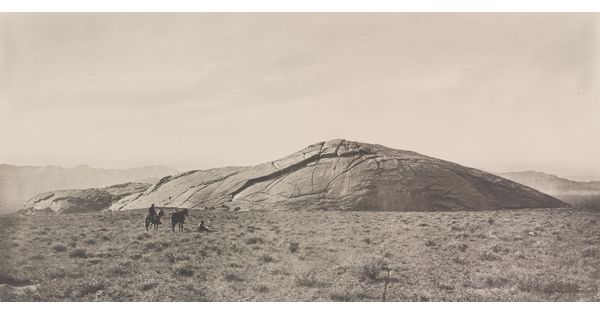 A black-and-white photograph of two people and two horses in a grassy field in front of a very large, smooth rock that seems to be emerging from the ground.