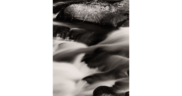 A black-and-white photograph of a water rushing over dark rocks.