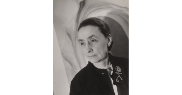 A black-and-white portrait photograph of an older White woman in front of a painting.