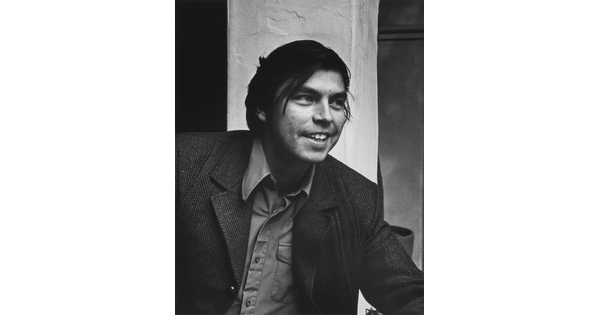 A black-and-white portrait photograph of a young medium-skinned man with longish dark hair, smiling, wearing a jacket over a button-down shirt.