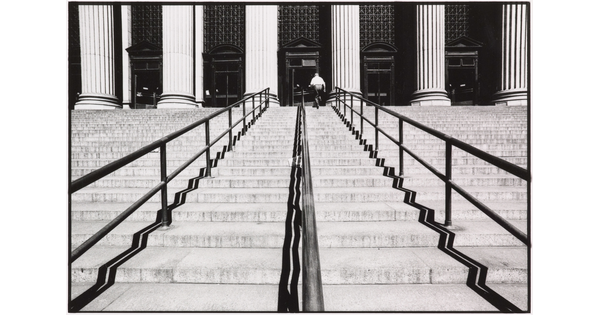 A black-and-white photograph of a person walking up a large set of stairs lined with handrails toward a building with large fluted columns.