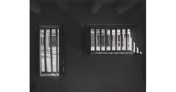 A black-and-white photograph taken from inside a dark room with a low ceiling looking toward a door and window with bars in them.