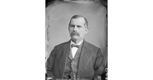 A black-and-white portrait image of a balding White man with a moustache wearing a dark three-piece suit with a bow tie.