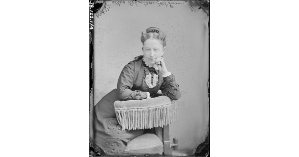 A black-and-white portrait image of a seated White woman, hair pulled to the top of her head, wearing a high-collared dress, leaning on the fringed arm of a chair, one hand raised to her cheek.