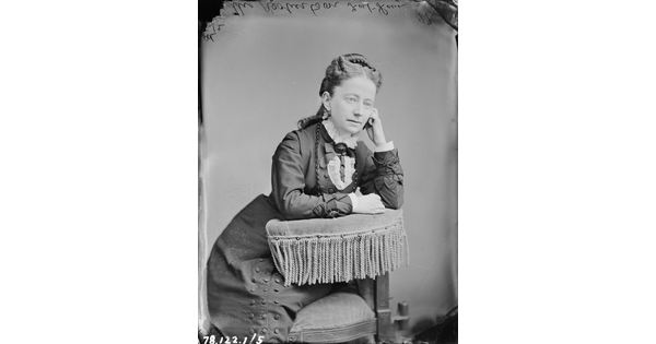 A black-and-white portrait image of a seated White woman, hair pulled to the top of her head, wearing a high-collared dress, leaning on the fringed arm of a chair, one hand raised to her cheek.