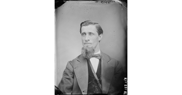A black-and-white portrait image of a seated White man with a long goatee wearing a bowtie, vest, and jacket.