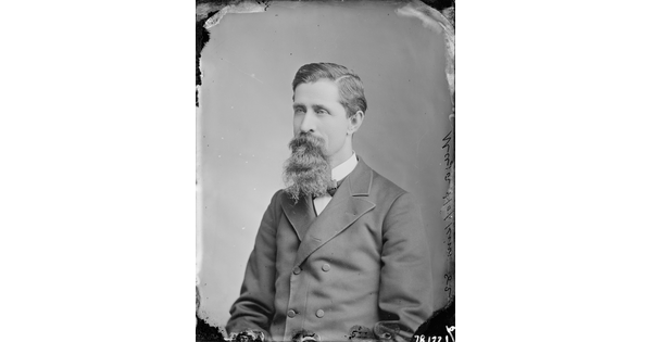 A black-and-white portrait image of a seated White man with a long mustache and goatee wearing a bowtie and double-breasted jacket.
