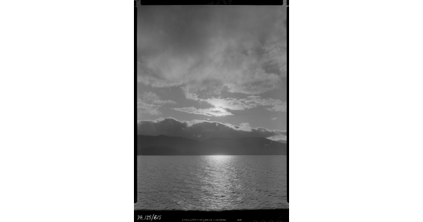 A black-and-white image of a lake, mountains on the opposite shore, under a cloudy sky at sunset.