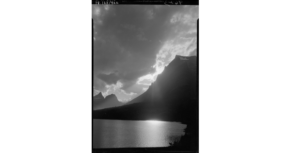A black-and-white image of a cloudy sky with the sun setting behind a mountain overlooking a still lake.