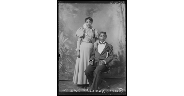A black-and-white studio image of a seated Black man in a dark suit and a standing Black woman wearing a long-sleeved full-length, light-colored dress resting one arm on the man's shoulder.