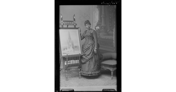 A black-and-white image of a Black woman wearing a long-sleeved Victorian-style dress standing next to an easel holding an artwork.