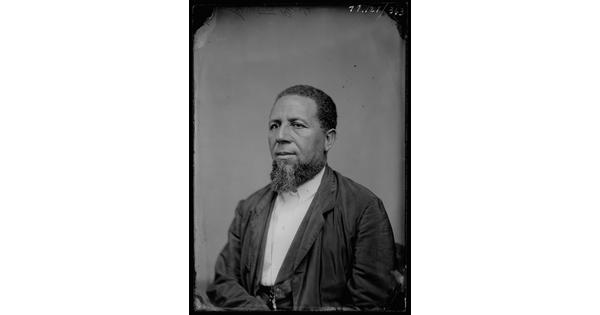 A black-and-white studio image of a seated Black man with a beard, wearing a white shirt and dark suit coat.