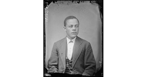 A black-and-white studio image of a seated young Black man with short dark hair, wearing a three-piece suit with a watch chain dangling from the vest.