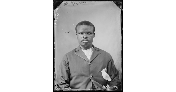 A black-and-white studio image of a seated young Black man wearing a suit jacket, a white pocket square stuffed into a breast pocket, over a white shirt.