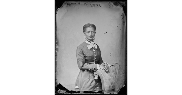A black-and-white studio image of a standing Black woman, hair in an updo, wearing a Victorian-style dress, hands folded over a chair.