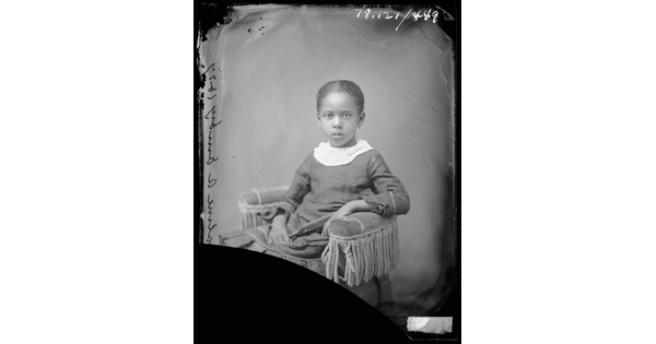 A black-and-white studio image of a seated Black girl wearing a dress with a lace collar.