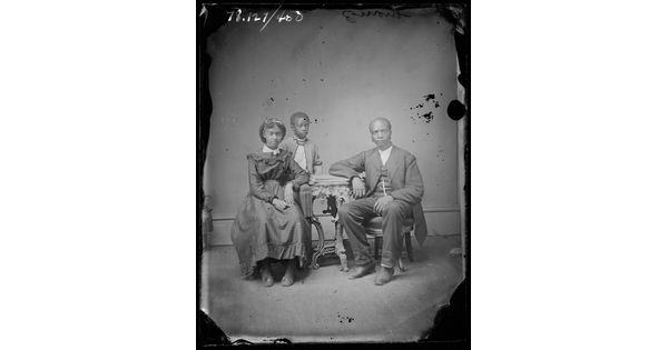A black-and-white portrait image of a Black family that shows a man and young woman sitting at a table and a child standing behind the woman.