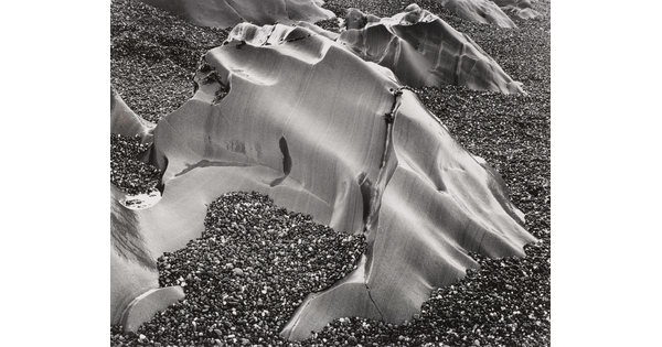 A black-and-white photograph of smooth, large, weather-worn rocks poking above a beach of smaller pebbles.