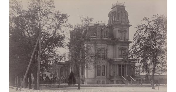 A black-and-white photograph of a four-story Victorian-style mansion with arched windows and a tower.