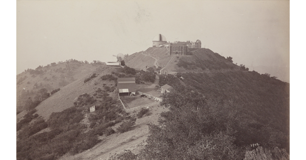 A sepia-toned photograph of a domed building and other structures atop a steep hillside covered with vegetation.