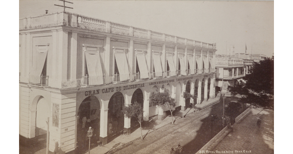 A black-and-white photograph of a white two-story hotel with arched entrances to an arcade on the street level.
