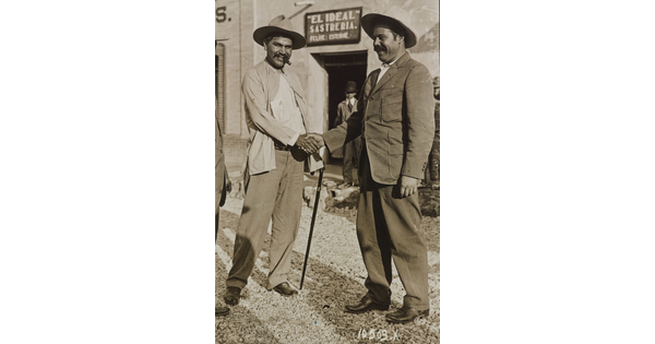 A black-and-white photograph of two medium-skinned men, both in suits and both wearing hats, shaking hands.