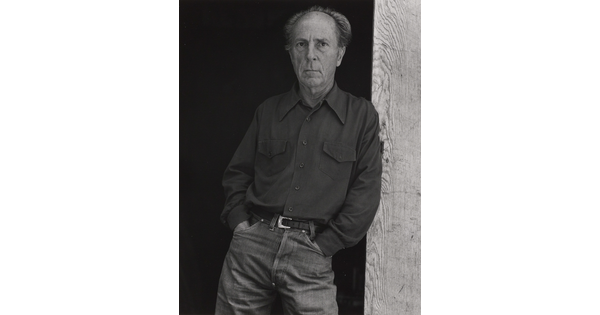 A black-and-white portrait of an older, balding White man wearing jeans and a long-sleeve button-down shirt leaning against a wooden beam.
