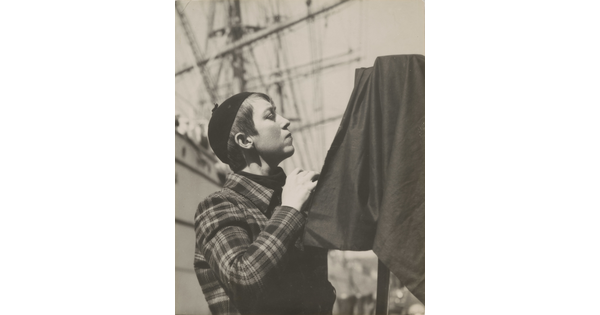 A black-and-white portrait photograph of a young, White woman in profile with short hair, wearing a beanie, standing behind a camera covered in black cloth.