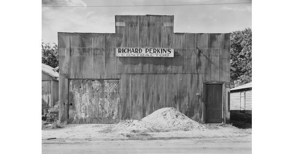 A black-and-white photograph of a building with a boarded up window and a pile of sand or gravel in front.