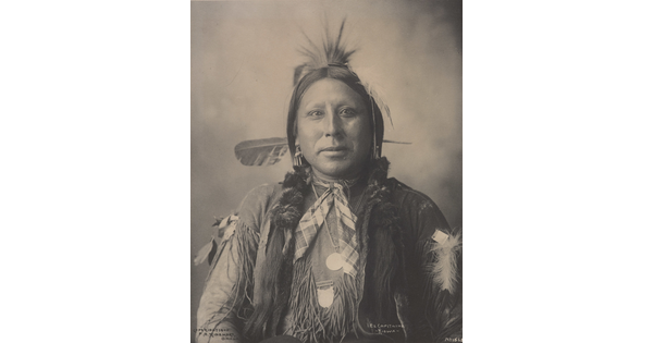 A black-and-white formal portrait of an Indigenous man in traditional clothing.